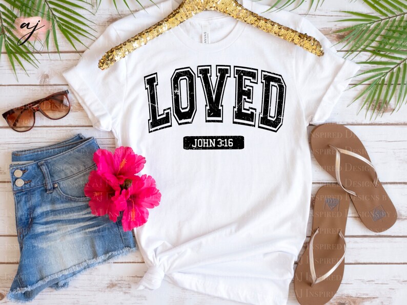 Op de afbeelding: Wit T-shirt met zwarte tekst die "LOVED" in een varsity-stijl lettertype leest. Onder de tekst staat "JOHN 3:16".