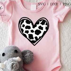 May include: A pink baby onesie with a black and white cow print heart design. The onesie is on a white surface with a gray stuffed elephant.
