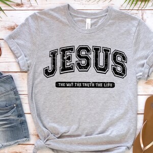 Op de afbeelding: Een lichtgrijs t-shirt met het woord "JESUS" in een zwarte, versleten lettertype in collegestijl. Daaronder staat de zin "THE WAY THE TRUTH THE LIFE" in een kleiner, zwart lettertype. Het t-shirt is opgevouwen en aan de onderkant geknoopt.