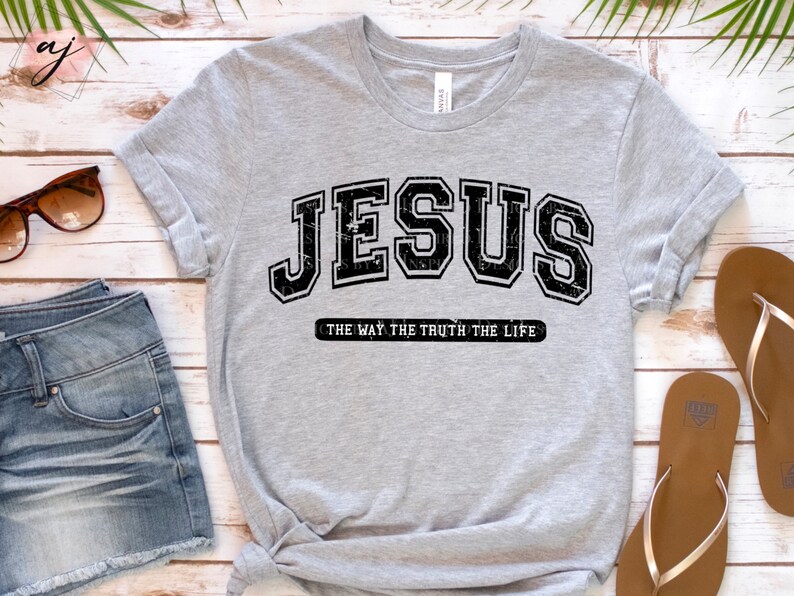 Op de afbeelding: Een lichtgrijs t-shirt met het woord "JESUS" in een zwarte, versleten lettertype in collegestijl. Daaronder staat de zin "THE WAY THE TRUTH THE LIFE" in een kleiner, zwart lettertype. Het t-shirt is opgevouwen en aan de onderkant geknoopt.