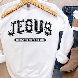 Op de afbeelding: Witte sweatshirt met het woord "JESUS" in een zwarte, versleten lettertype in collegestijl. Daaronder staat de tekst "THE WAY THE TRUTH THE LIFE" in het zwart. De sweatshirt wordt getoond met een blauwe jeans en bruine enkellaarsjes.