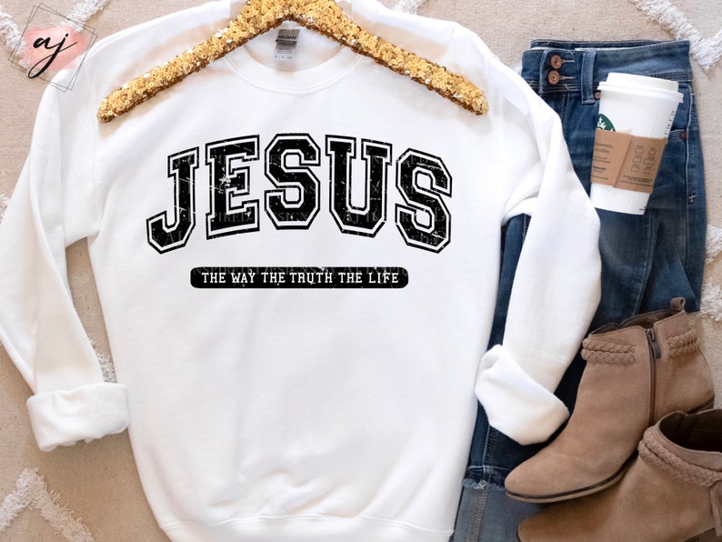 Op de afbeelding: Witte sweatshirt met het woord "JESUS" in een zwarte, versleten lettertype in collegestijl. Daaronder staat de tekst "THE WAY THE TRUTH THE LIFE" in het zwart. De sweatshirt wordt getoond met een blauwe jeans en bruine enkellaarsjes.