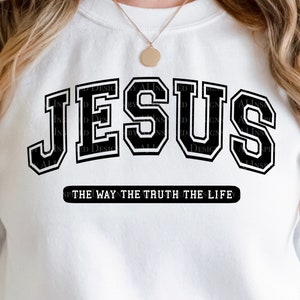 Op de afbeelding: Witte sweatshirt met het woord "JESUS" in grote, zwarte letters in collegestijl. Daaronder staat de tekst "THE WAY THE TRUTH THE LIFE". Een eenvoudig, casual kledingstuk.