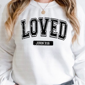 Op de afbeelding: Wit sweatshirt met zwarte tekst die "LOVED" luidt in een varsity-stijl lettertype. Onder de tekst staat "JOHN 3:16" in een kleiner lettertype.