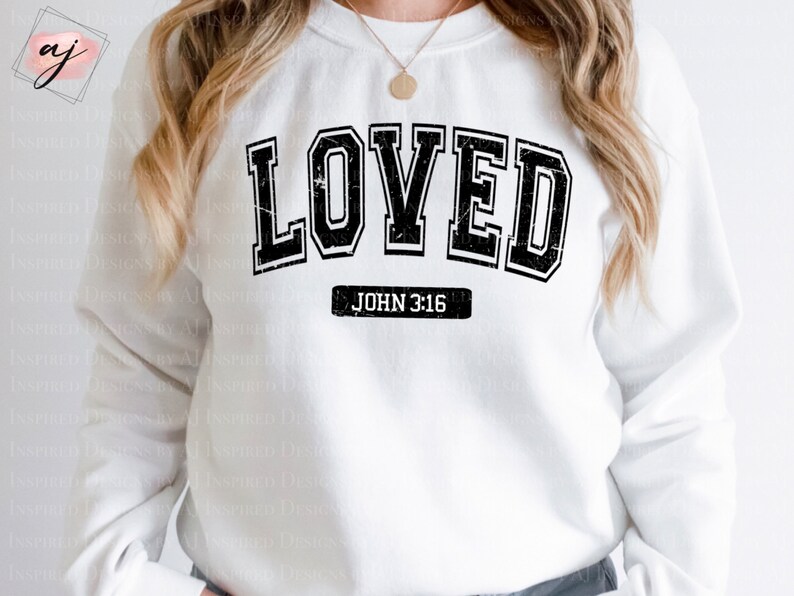 Op de afbeelding: Wit sweatshirt met zwarte tekst die "LOVED" luidt in een varsity-stijl lettertype. Onder de tekst staat "JOHN 3:16" in een kleiner lettertype.