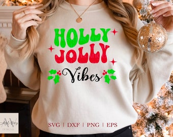 May include: A beige sweatshirt with the words "HOLLY JOLLY Vibes" in green, red, and black. The design includes holly leaves and berries, and red star accents. The sweatshirt is a festive holiday apparel item.