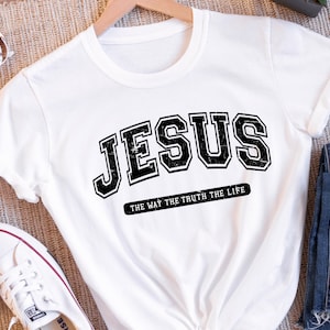 Op de afbeelding: Wit T-shirt met het woord "JESUS" in een zwarte, verweerde letter in collegestijl. Daaronder staat de zin "THE WAY THE TRUTH THE LIFE" in kleinere, zwarte letters. Het T-shirt is opgevouwen.