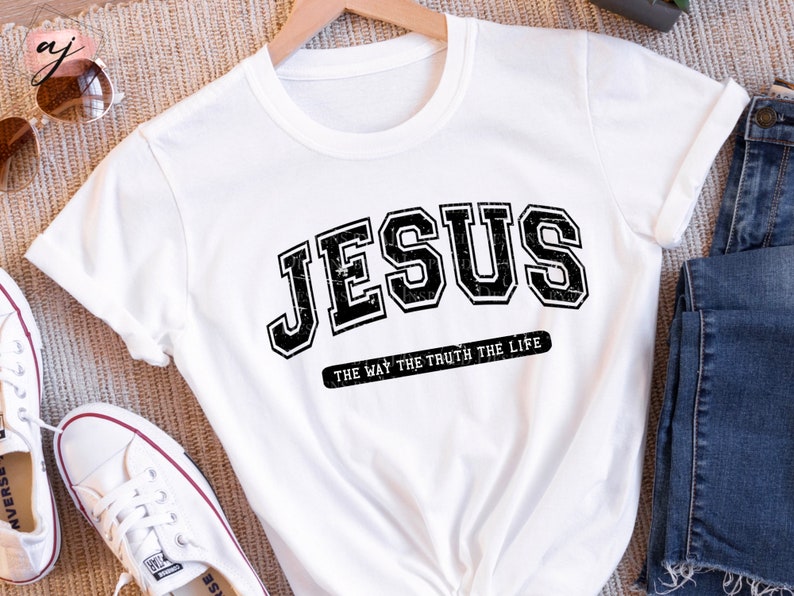 Op de afbeelding: Wit T-shirt met het woord "JESUS" in een zwarte, verweerde letter in collegestijl. Daaronder staat de zin "THE WAY THE TRUTH THE LIFE" in kleinere, zwarte letters. Het T-shirt is opgevouwen.