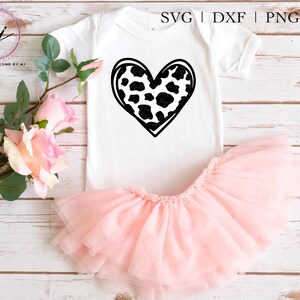 May include: A white onesie with a black and white cow print heart design. The onesie is paired with a pink tulle tutu.