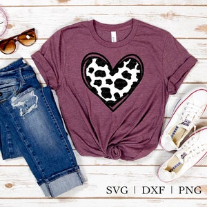 May include: A maroon t-shirt with a black and white cow print heart design. The shirt is laid flat on a white wooden surface with a pair of sunglasses, a pair of jeans, and a pair of white sneakers.