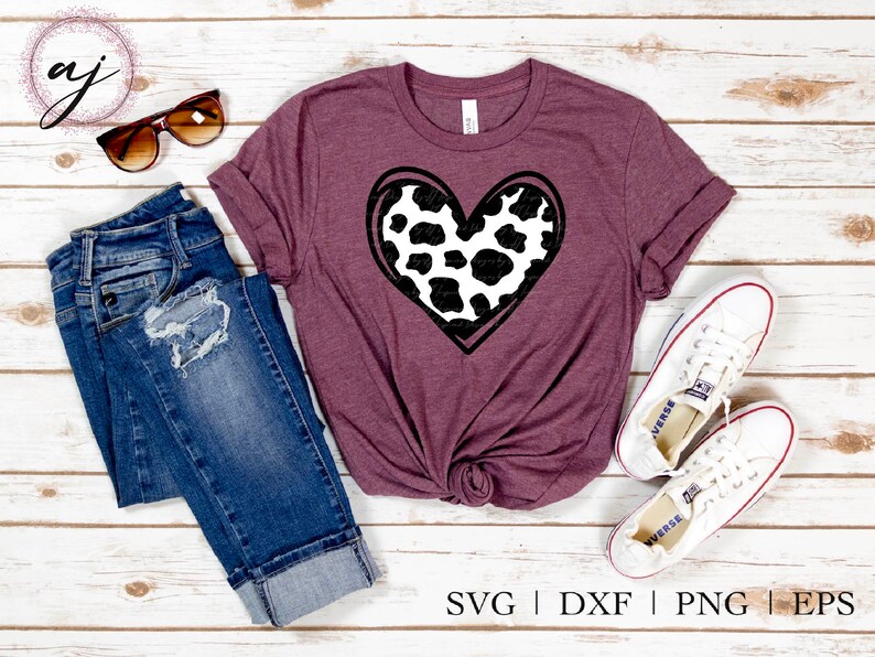 May include: A maroon t-shirt with a black and white cow print heart design. The shirt is laid flat on a white wooden surface with a pair of sunglasses, a pair of jeans, and a pair of white sneakers.