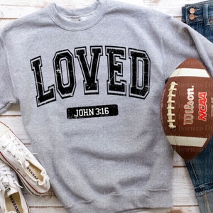 Op de afbeelding: Grijs sweatshirt met het woord "LOVED" in zwarte blokletters met een verweerde look. De tekst "JOHN 3:16" is onder het woord "LOVED" gedrukt.