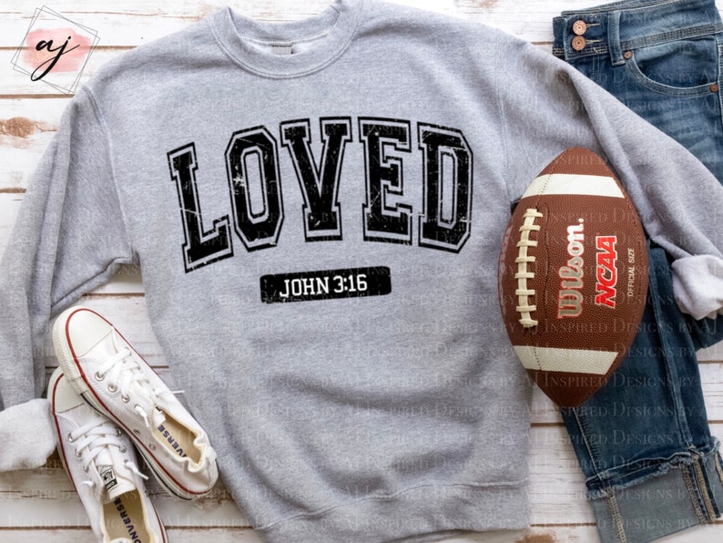 Op de afbeelding: Grijs sweatshirt met het woord "LOVED" in zwarte blokletters met een verweerde look. De tekst "JOHN 3:16" is onder het woord "LOVED" gedrukt.
