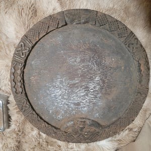 Puede incluir: Una bandeja circular de madera de color marrón oscuro con un centro liso y plano, y un borde ancho y elevado decorado con intrincados patrones geométricos tallados a mano, incluyendo rayado cruzado, surcos lineales y símbolos estilizados.