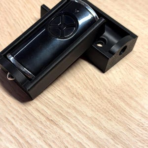 May include: A black garage door opener remote control with silver trim, in a black holder. The remote features a circular button design. The holder is open, showing the remote.