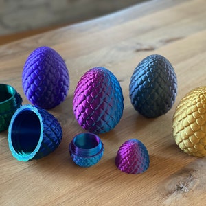 Könnte beinhalten: Sechs dracheneierförmige Schmuckkästchen, einige geöffnet, in verschiedenen Metallic-Farben, darunter Gold, Blau, Grün und Lila. Die Kästchen sind mit einer schuppigen Textur verziert.