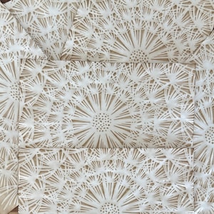 May include: Close-up of a fabric with a repeating dandelion pattern. The dandelions are white, with intricate details, set against a tan background. The fabric is folded, showing multiple panels.