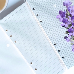 May include: A white notebook with lined and gridded pages. The notebook has holes on the side for a binder. A sprig of purple lavender flowers is in the bottom right corner of the image.