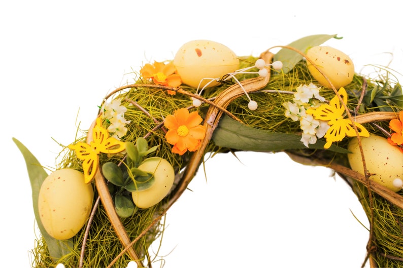 K&ouml;nnte beinhalten: Ein gr&uuml;ner und brauner Osterkranz, der mit gelben und orangefarbenen Blumen, gelben Schmetterlingen und gelben Eiern verziert ist.