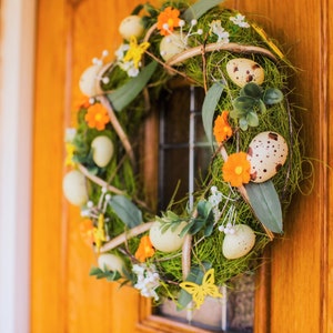 K&ouml;nnte beinhalten: Ein gr&uuml;ner und brauner Kranz mit orangefarbenen Blumen und gefleckten Eiern. Der Kranz h&auml;ngt an einer Holzt&uuml;r.