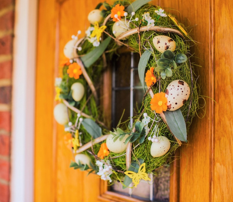 K&ouml;nnte beinhalten: Ein gr&uuml;ner und brauner Kranz mit orangefarbenen Blumen und gefleckten Eiern. Der Kranz h&auml;ngt an einer Holzt&uuml;r.