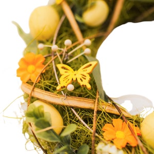 K&ouml;nnte beinhalten: Ein gr&uuml;ner Osterkranz mit gelben Eiern, orangefarbenen Blumen und einem gelben Schmetterling. Der Kranz ist aus Zweigen gefertigt und hat einen rustikalen Look.