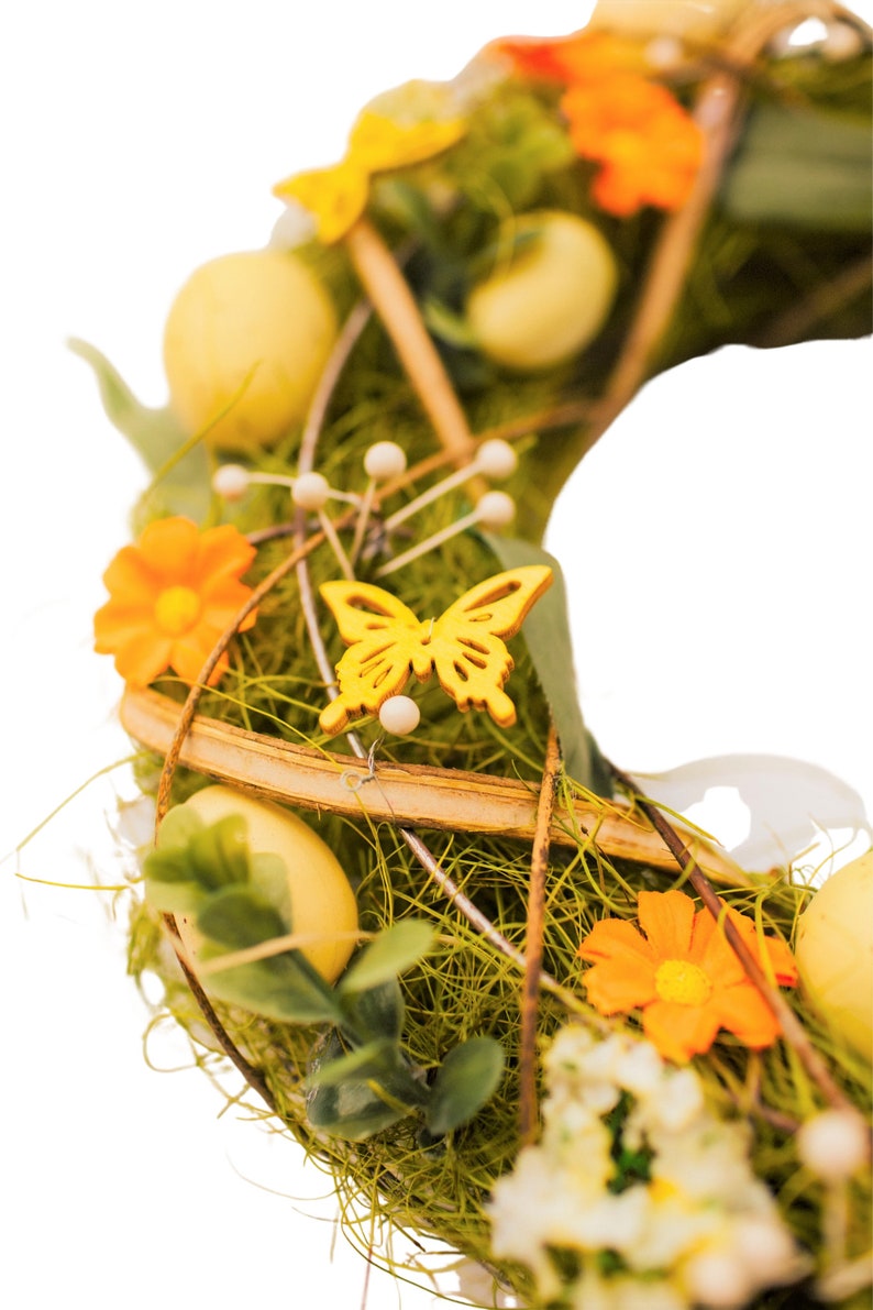 K&ouml;nnte beinhalten: Ein gr&uuml;ner Osterkranz mit gelben Eiern, orangefarbenen Blumen und einem gelben Schmetterling. Der Kranz ist aus Zweigen gefertigt und hat einen rustikalen Look.