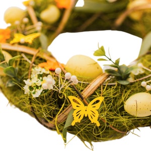 K&ouml;nnte beinhalten: Ein gr&uuml;ner Osterkranz mit gelben Eiern, orangefarbenen Blumen und gelben Schmetterlingen. Der Kranz ist aus Zweigen und Moos gefertigt.