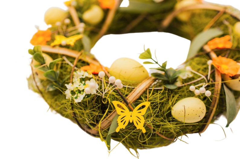 K&ouml;nnte beinhalten: Ein gr&uuml;ner Osterkranz mit gelben Eiern, orangefarbenen Blumen und gelben Schmetterlingen. Der Kranz ist aus Zweigen und Moos gefertigt.