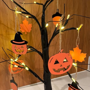 Op de afbeelding: Een zwarte takkenboom met oranje en zwarte Halloween-decoraties. De boom is versierd met pompoenen, bladeren en eikels. De boom is verlicht met warmwitte lampjes.
