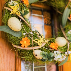 K&ouml;nnte beinhalten: Ein gr&uuml;ner Fr&uuml;hlingskranz mit gelben Schmetterlingen, orangefarbenen Blumen und wei&szlig;en Eiern. Der Kranz h&auml;ngt an einer Holzt&uuml;r.