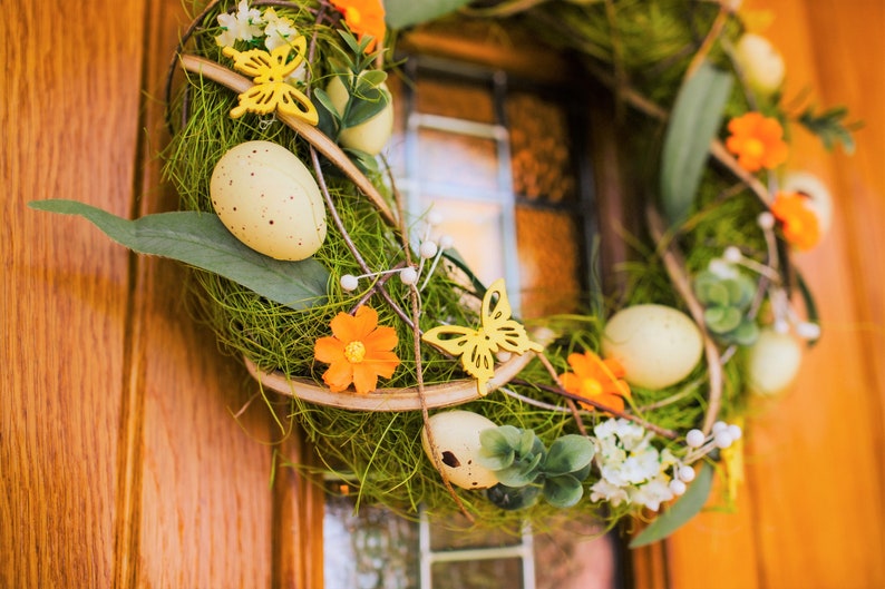 K&ouml;nnte beinhalten: Ein gr&uuml;ner Fr&uuml;hlingskranz mit gelben Schmetterlingen, orangefarbenen Blumen und wei&szlig;en Eiern. Der Kranz h&auml;ngt an einer Holzt&uuml;r.