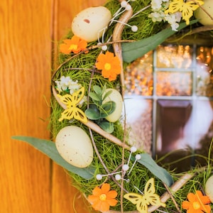 K&ouml;nnte beinhalten: Ein Fr&uuml;hlingskranz mit gr&uuml;nem Laub, wei&szlig;en und gelben Bl&uuml;ten und gelben Schmetterlingen. Der Kranz ist mit wei&szlig;en, gefleckten Eiern verziert.