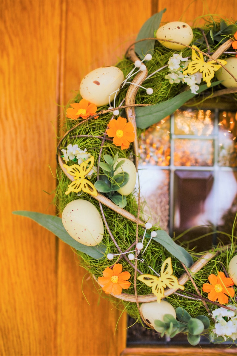 K&ouml;nnte beinhalten: Ein Fr&uuml;hlingskranz mit gr&uuml;nem Laub, wei&szlig;en und gelben Bl&uuml;ten und gelben Schmetterlingen. Der Kranz ist mit wei&szlig;en, gefleckten Eiern verziert.