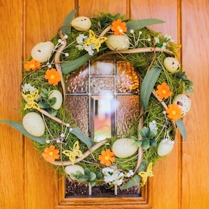 K&ouml;nnte beinhalten: Ein gr&uuml;ner und brauner Osterkranz mit gelben Schmetterlingen, orangefarbenen Blumen und wei&szlig;en, gefleckten Eiern.