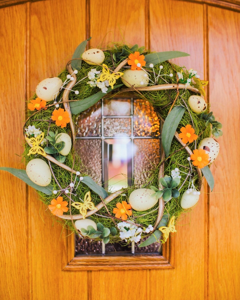K&ouml;nnte beinhalten: Ein gr&uuml;ner und brauner Osterkranz mit gelben Schmetterlingen, orangefarbenen Blumen und wei&szlig;en, gefleckten Eiern.