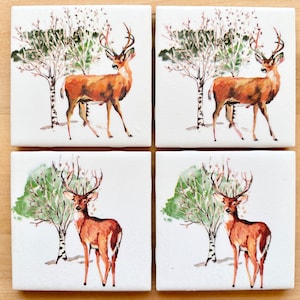 Op de afbeelding: Set van vier keramische onderzetters met een aquarel-illustratie van een hert dat voor een boom staat. De onderzetters zijn wit met een lichtbruine achtergrond en groen gebladerte.