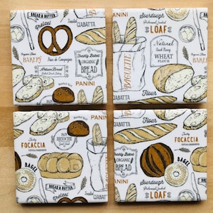 Op de afbeelding: Set van zes witte keramische onderzetters met bruine en gouden illustraties van verschillende soorten brood, waaronder zuurdesem, baguette, focaccia en pretzels. De onderzetters bevatten tekst zoals "Sourdough", "Baguette", "Focaccia", "Pretzel", "Pain de Campagne", "Pain Brioche", "Biologisch brood", "Tarwemeel", "Panini" en "Ciabatta".