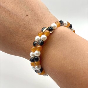 Könnte beinhalten: Ein mehrreihiges Perlenarmband mit einem sich wiederholenden Muster aus glatten weißen Kugeln, durchscheinenden bernsteinfarbenen Perlen und klaren Perlen mit schwarzen Einschlüssen, das in einem geschichteten Design um das Handgelenk liegt.