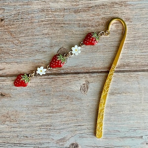 May include: A gold-colored bookmark with a decorative chain of red strawberries and white flowers. The bookmark has a curved top and a floral pattern on the lower portion. The strawberries have green leaves.