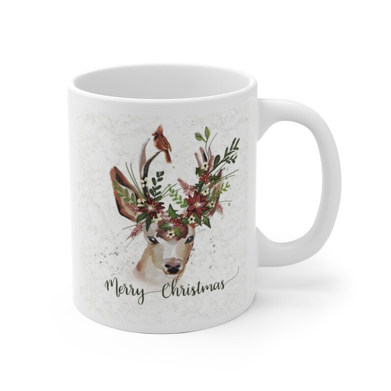 May include: White ceramic mug with a watercolor illustration of a deer with a floral crown and a cardinal bird perched on its antlers. The text "Merry Christmas" is written below the deer.