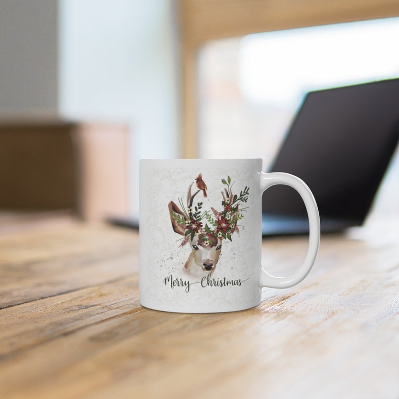 May include: White ceramic mug with a floral wreath and a deer head design. The mug has the text "Merry Christmas" on the front.
