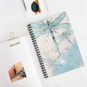 Puede incluir: Un cuaderno de espiral con un diseño de acuarela de tres libélulas. Las libélulas son azules y blancas y tienen alas translúcidas. El fondo es una lavada de acuarela azul claro.