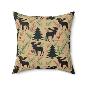 Puede incluir: Una almohada beige con un patrón repetido de alces, pinos y hojas. Los alces son siluetas negras y los árboles son verdes. El texto "I love Moose" se repite en todo el patrón.
