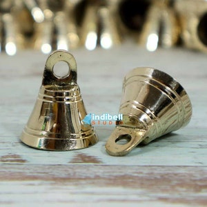 Puede incluir: Dos pequeñas campanas doradas con la parte superior redondeada y un pequeño lazo en la parte superior. Las campanas están sobre una superficie de madera clara. Hay muchas otras campanas en el fondo.