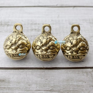 May include: Three gold-colored metal bells with a detailed lion face design. The bells have a loop at the top for hanging.