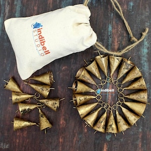 May include: A set of 20 small, gold-coloured metal bells with a rustic finish. The bells are strung together in a circle and come in a small white drawstring bag with the logo "indibell studio".