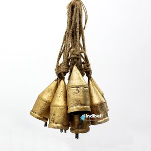 May include: A cluster of seven antique-style brass bells suspended from a natural jute rope. The bells have a conical shape with a slightly aged, golden finish. The image is set against a plain white background.