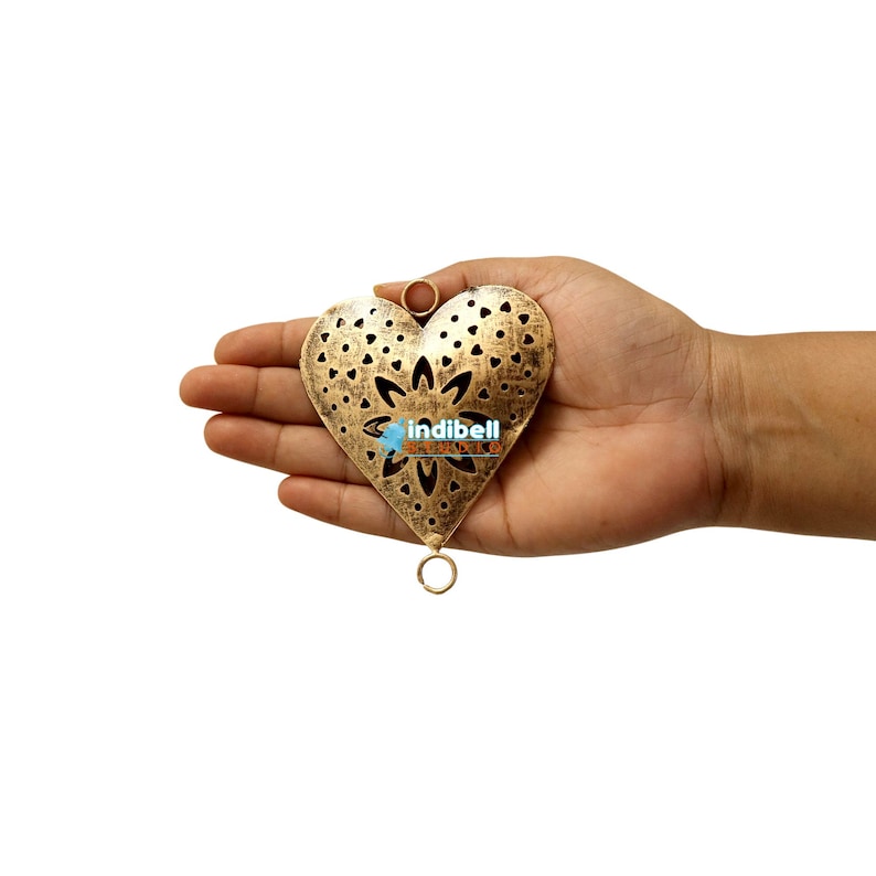A hand displaying a heart-shaped metal cutout with rustic vintage detailing and a floral pattern.