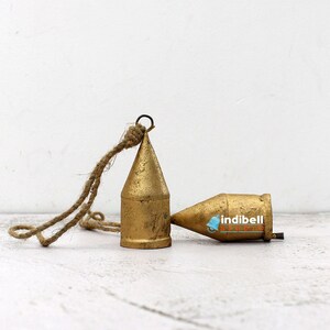 May include: Two antique brass-colored bells with a rustic rope hanging from one. The bells have a conical shape and a weathered finish, suggesting age and use. The bells are resting on a white surface.